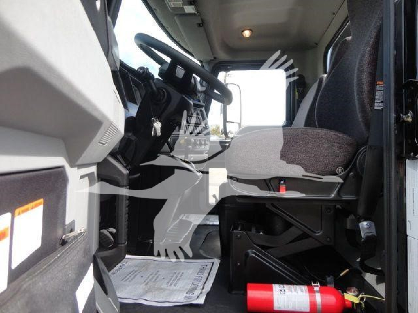 Truck cab interior view of a 2026 Mack MD6 showing driver's seat, steering wheel, and pedal area with a fire extinguisher mounted under the dash.