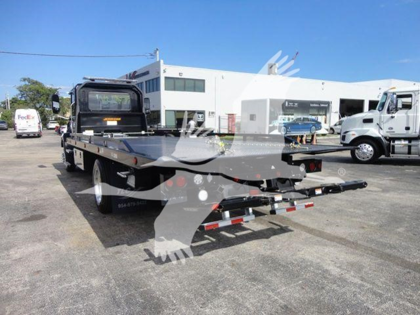 Flatbed rollback tow truck trailer in a dealership lot with a Mack MD6 in background, rear view showing dual ramps and deck.