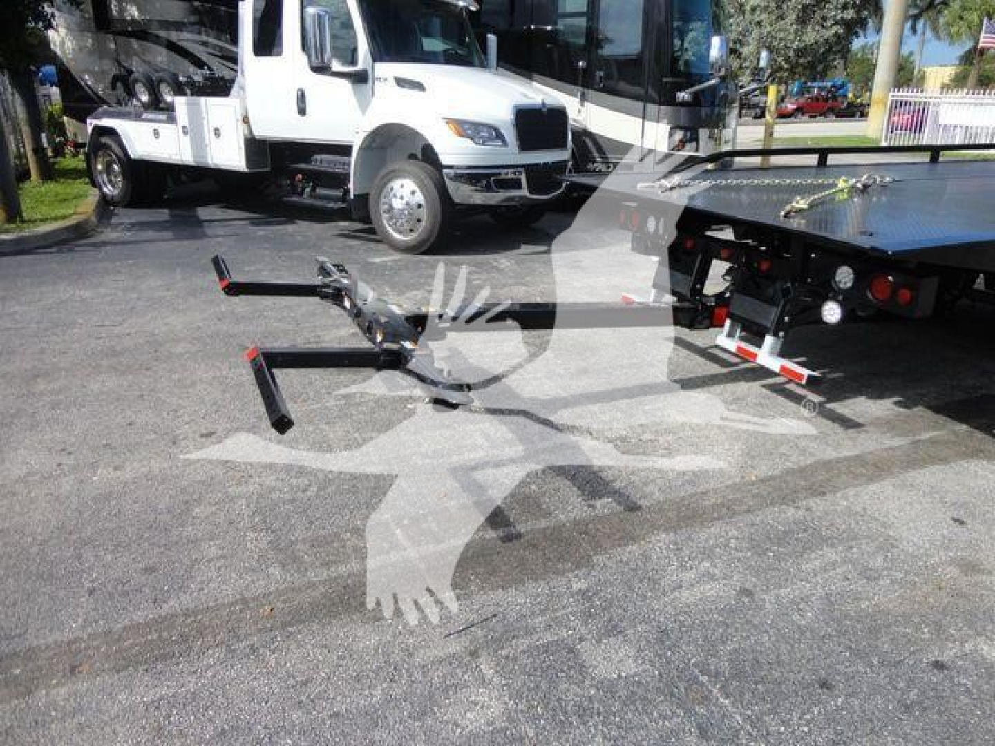 Trucker’s platform with a Mack MD6 rollback truck in background; front hitch and tow arms extended in foreground; white vehicle and parking lot.