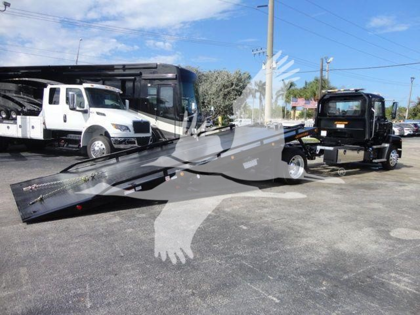 Truck delivery scene with a rollback bed trailer in foreground and a white tow truck and a black tractor unit in background; flat asphalt lot under blue sky.