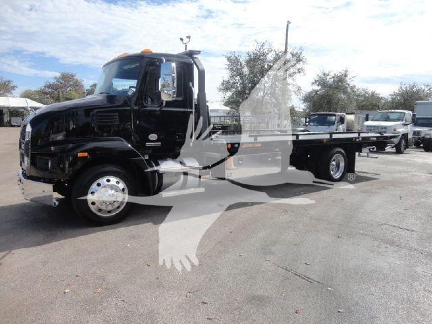 MD6 rollback truck with black cab and flatbed in-service condition, Mack badge visible, side mirrors, orange clearance lamps, distant equipment in lot.