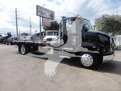 2026 Mack MD6 rollback truck showing black cab, chrome bumper, flatbed with stake pockets, and wheel covers. visible text on signs in background blurred.
