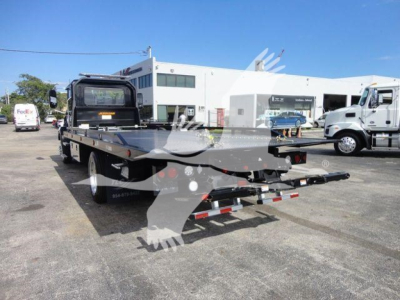 Flatbed rollback tow truck trailer in a dealership lot with a Mack MD6 in background, rear view showing dual ramps and deck.
