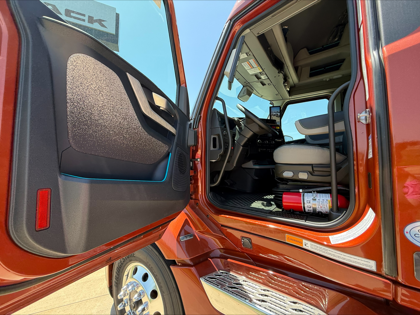  Volvo VNL cabin interior view with open driver door showing steering wheel, dashboard, and passenger seat. Fire extinguisher visible on floor near door.