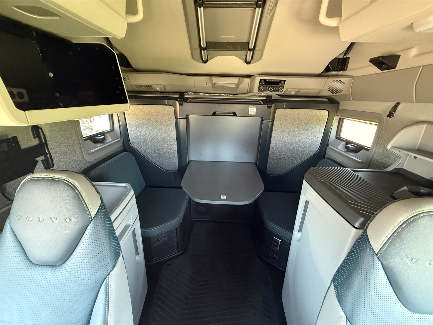 Truck cab interior with seating and table in sleeper area. Volvo branding on seats. Overhead bunk area with storage. Windows on both sides.