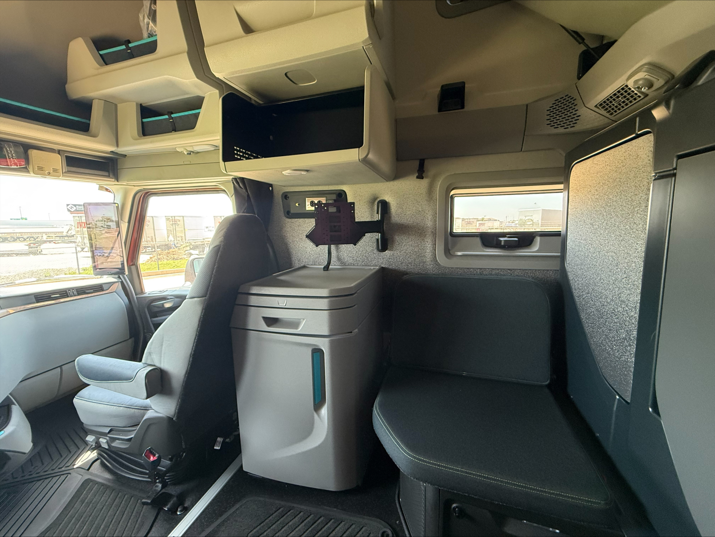 Interior cab of a 2026 Volvo VNL64T860 sleeper truck showing driver's seat, overhead storage, side window, and a compact cabinet with a freezer/refrigerator unit.