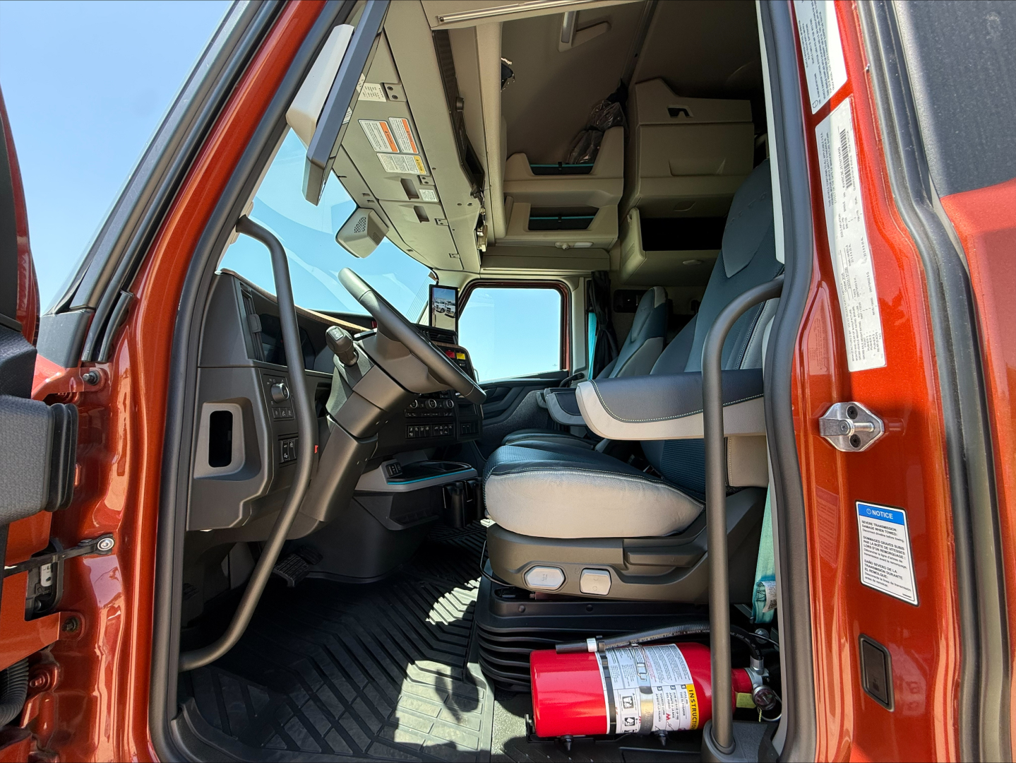 Interior view of a Volvo VNL64T860 truck cab from the driver’s side door open, showing dash, steering wheel, seat, overhead storage, and a red fire extinguisher on the floor.