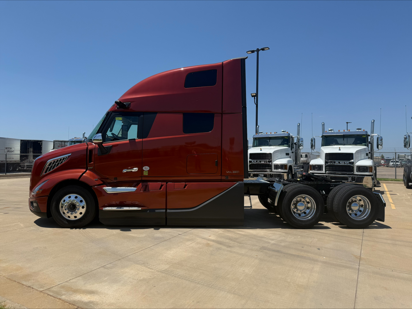 Volvo VNL64T860 truck side view showing red cab with sleeper, dual rear axles, chrome accents, and white trucks in background.