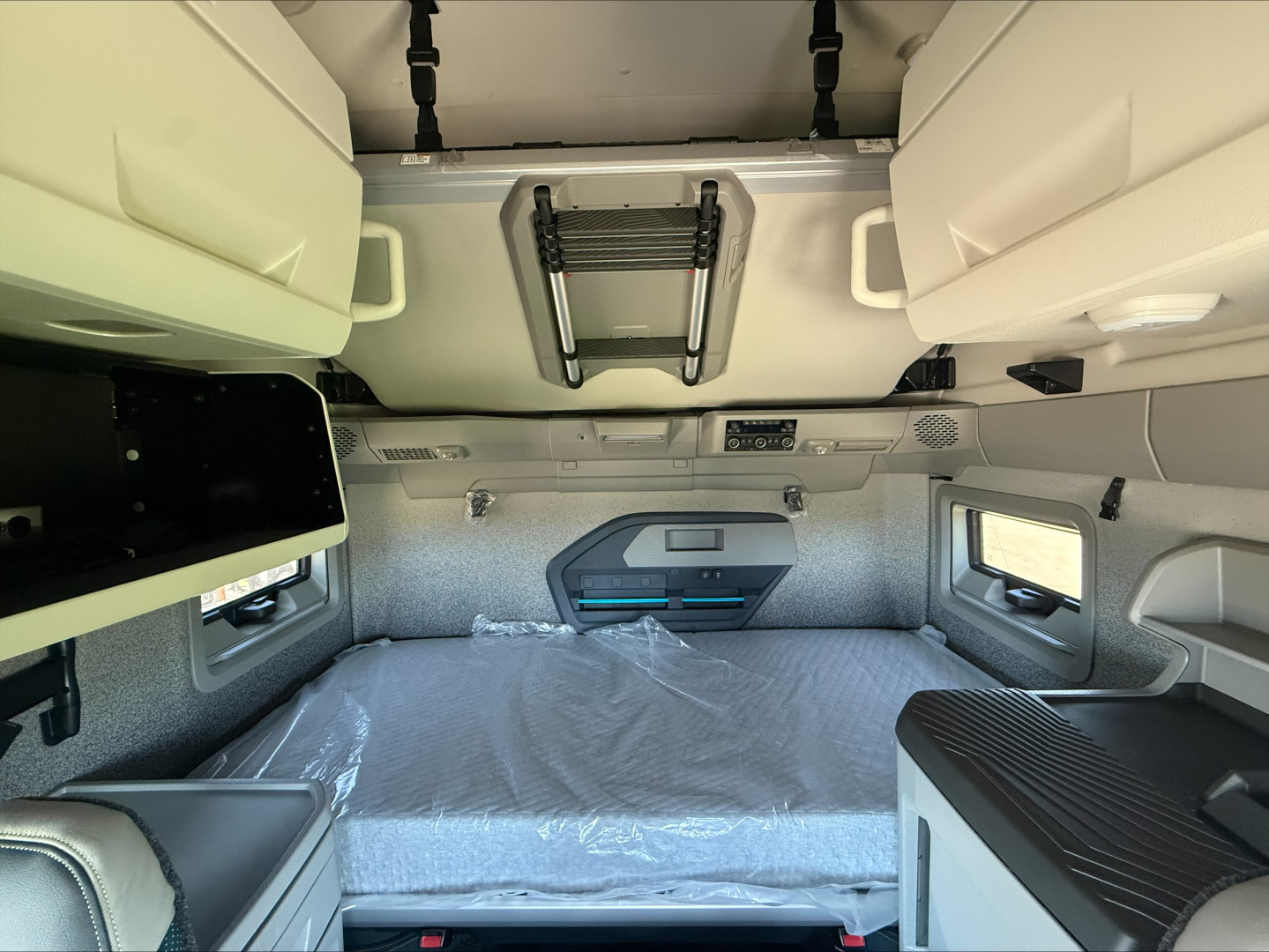 Volvo VNL64T860 sleeper cab interior showing bed area with plastic covering, overhead storage bins, ladder rack, window, and side cabinetry.