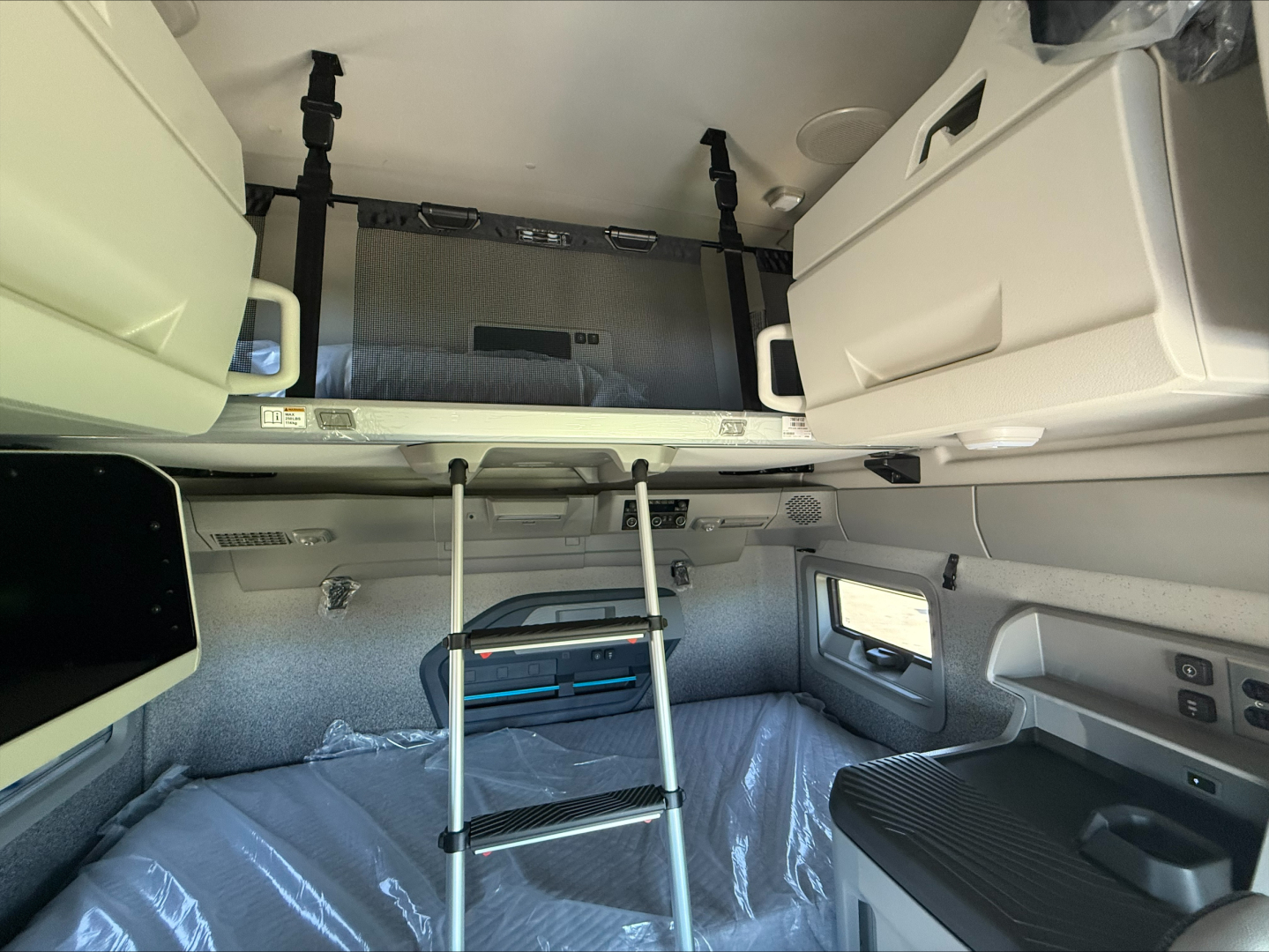 Interior of a Volvo VNL sleeper cab showing upper storage bunk, ladder, and window; includes white cabinets and grey upholstery.