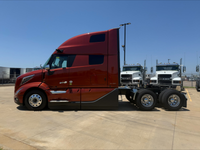 Volvo VNL64T860 truck side view showing red cab with sleeper, dual rear axles, chrome accents, and white trucks in background.