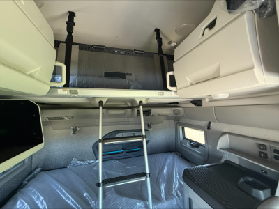 Interior of a Volvo VNL sleeper cab showing upper storage bunk, ladder, and window; includes white cabinets and grey upholstery.
