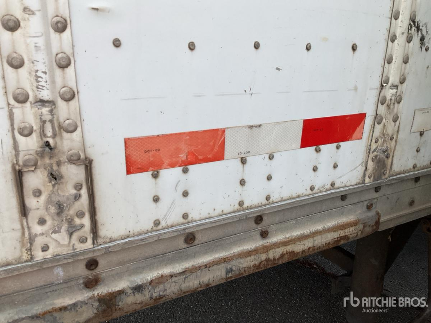 Trailer side with riveted metal panels, white paint, and red/white reflective strip; rust on lower edge and frame visible.