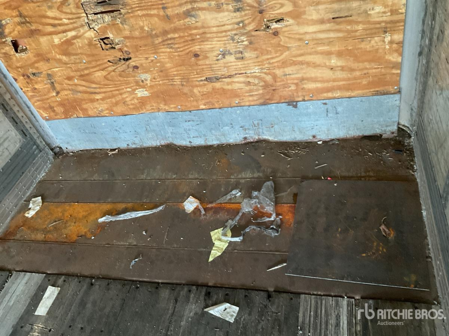 Trailer interior with wooden and metal floor, debris, and worn plywood wall. Visible text includes no legible labels; overall a rough cargo area.