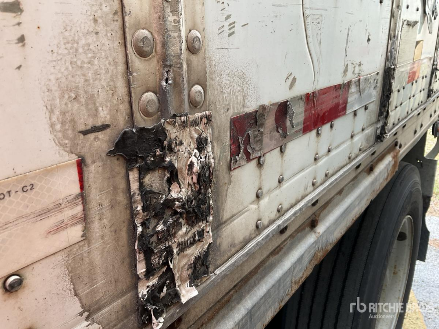 Trailer side with damaged side panel and riveted metal; visible burnt or charred area around a damaged patch near door hinge; no legible alphanumeric text clearly visible in this crop.
