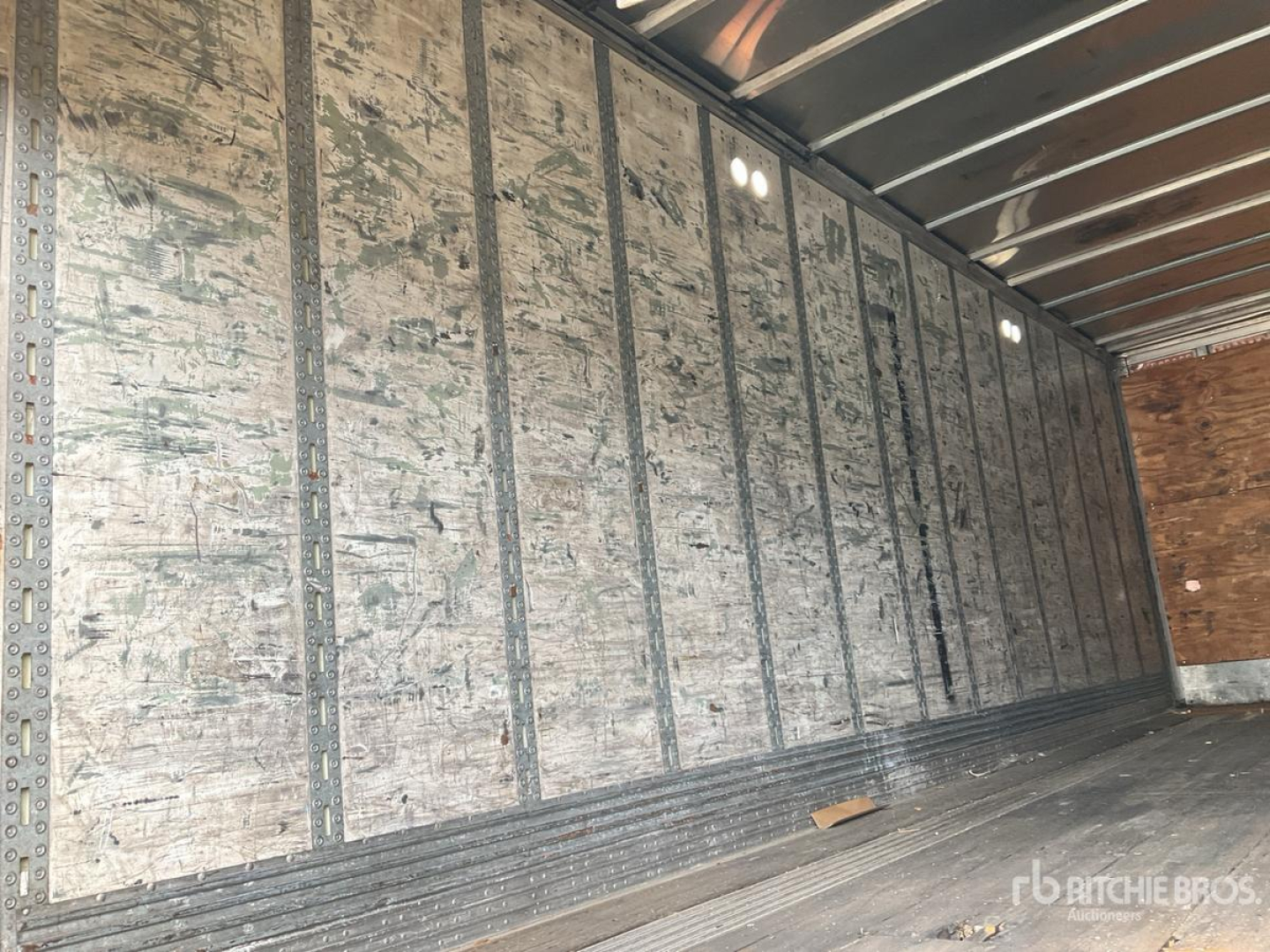Trailer interior with silver metal walls and ribbed floor showing worn plywood header; no text readable.