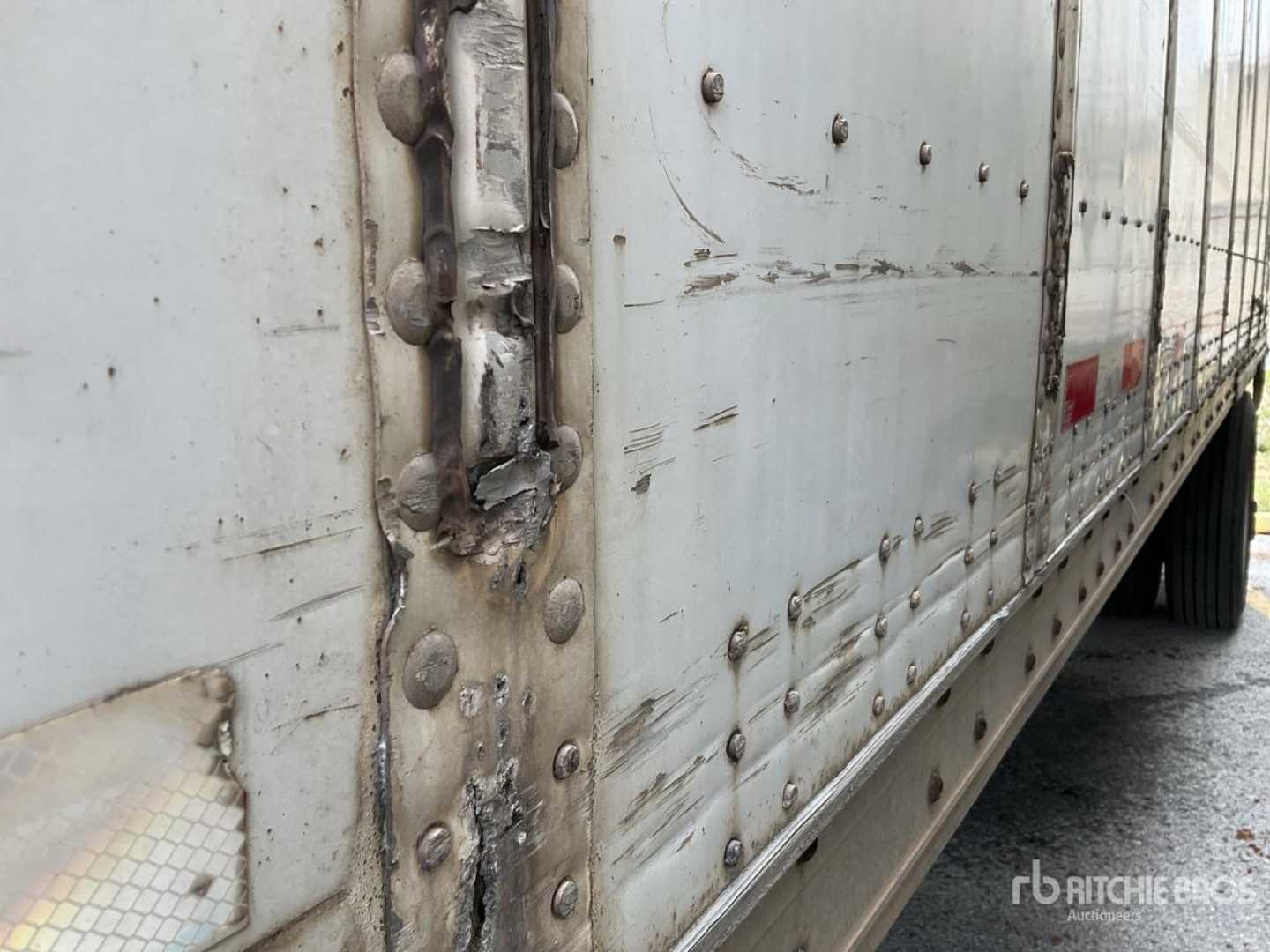 Trailer side view showing riveted aluminum skin, vertical seam, and welded longitudinal rail with protective trim; damaged area near rear door hinge.