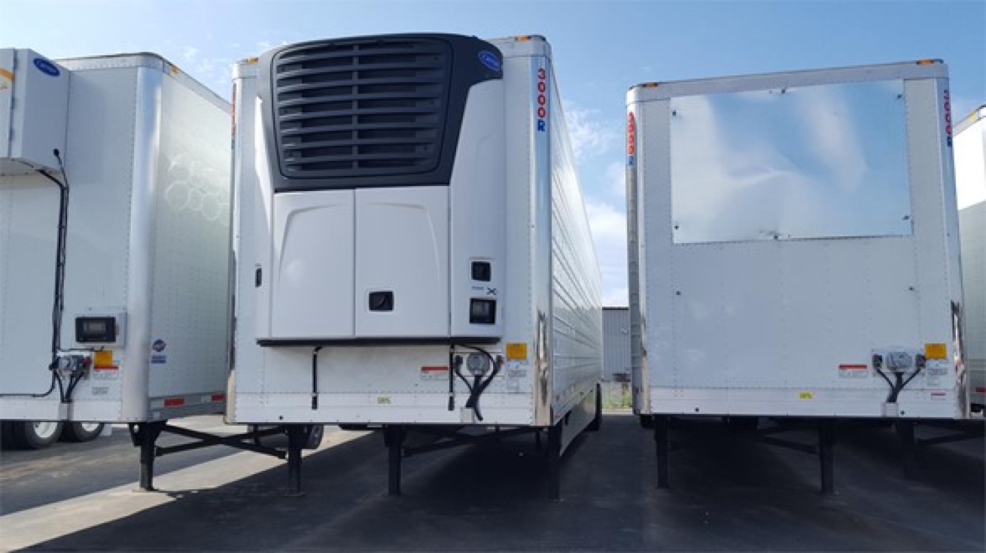 Front view of a white refrigerated trailer unit with a large black front grille and cooling unit, flanked by two silver trailers, on a lot.