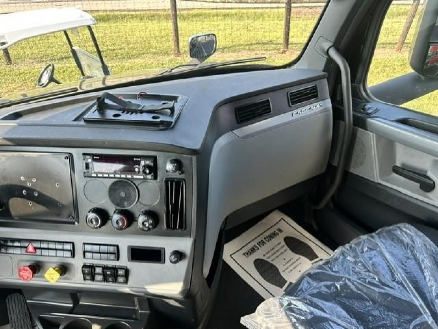 Cab interior of a Freightliner Cascadia 126 showing dashboard with steering wheel, vents, radio, and plastic protective seat cover.