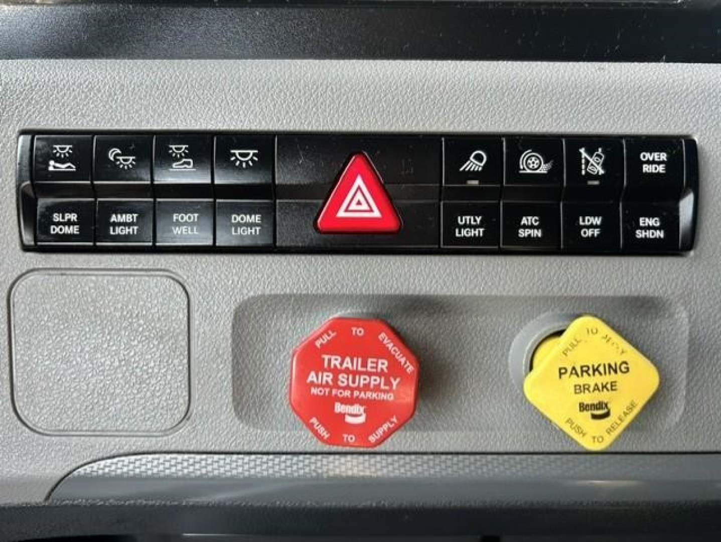 Dashboard control panel of a 2022 Freightliner Cascadia 126 showing hazard button, auxiliary switches, red trailer air supply cap, and yellow parking brake cap.