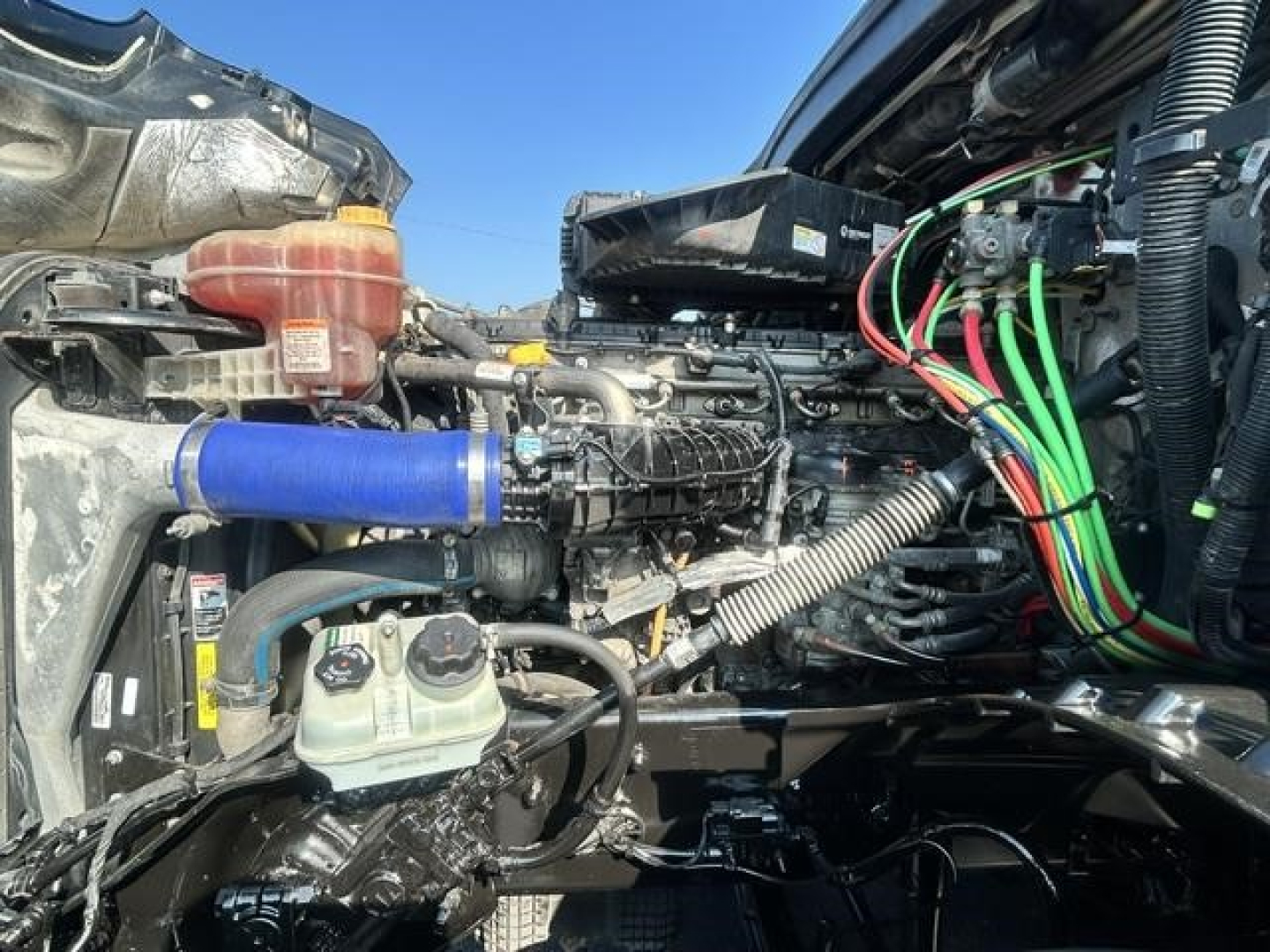 Truck engine bay showing coolant reservoir, hoses, electrical loom, and turbo/intercooler piping in a Freightliner Cascadia engine compartment.