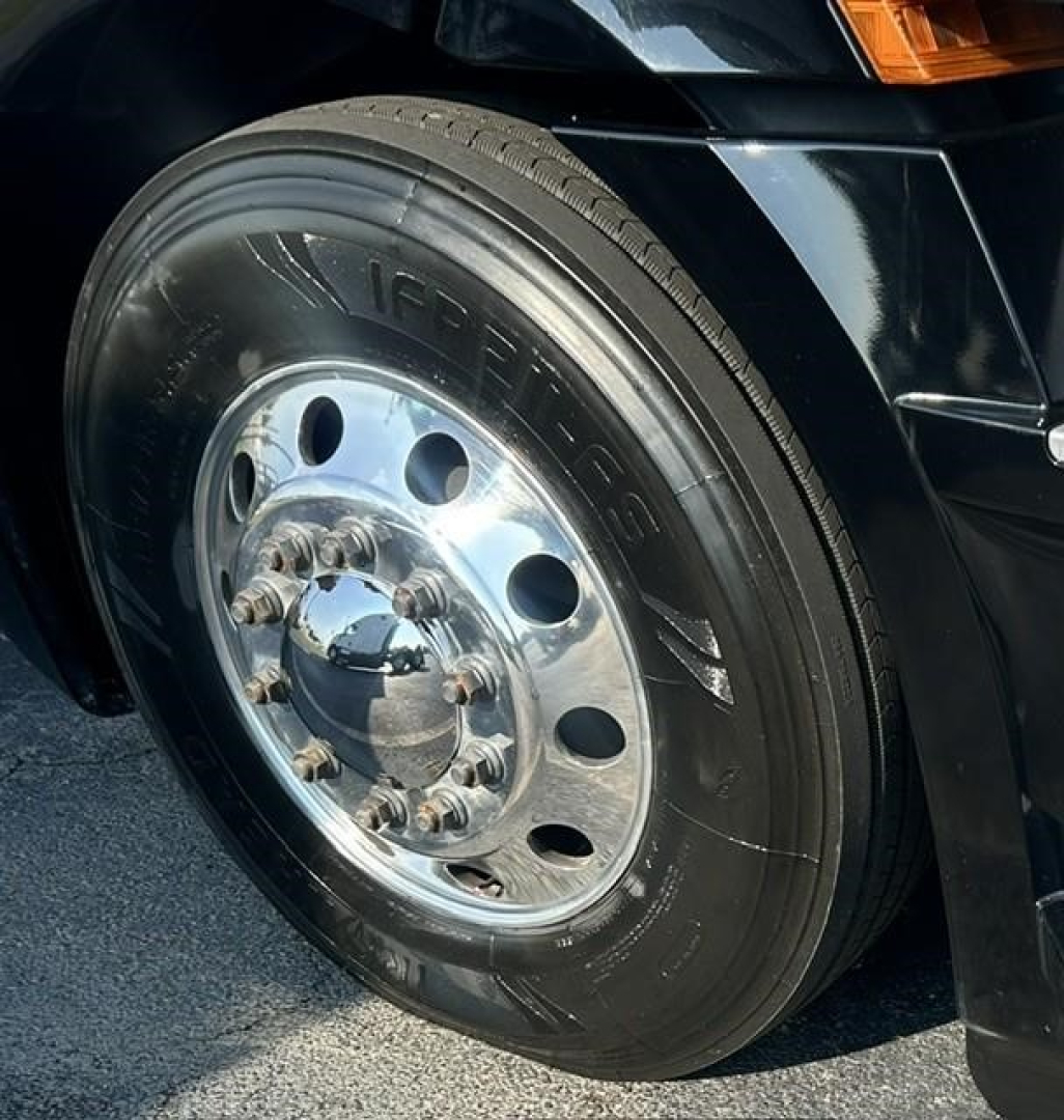Truck wheel and tire close-up on a Freightliner Cascadia 126. Visible: polished chrome wheel hub, lug nuts, tire with brand marking IR something, fender and headlight area in black paint.