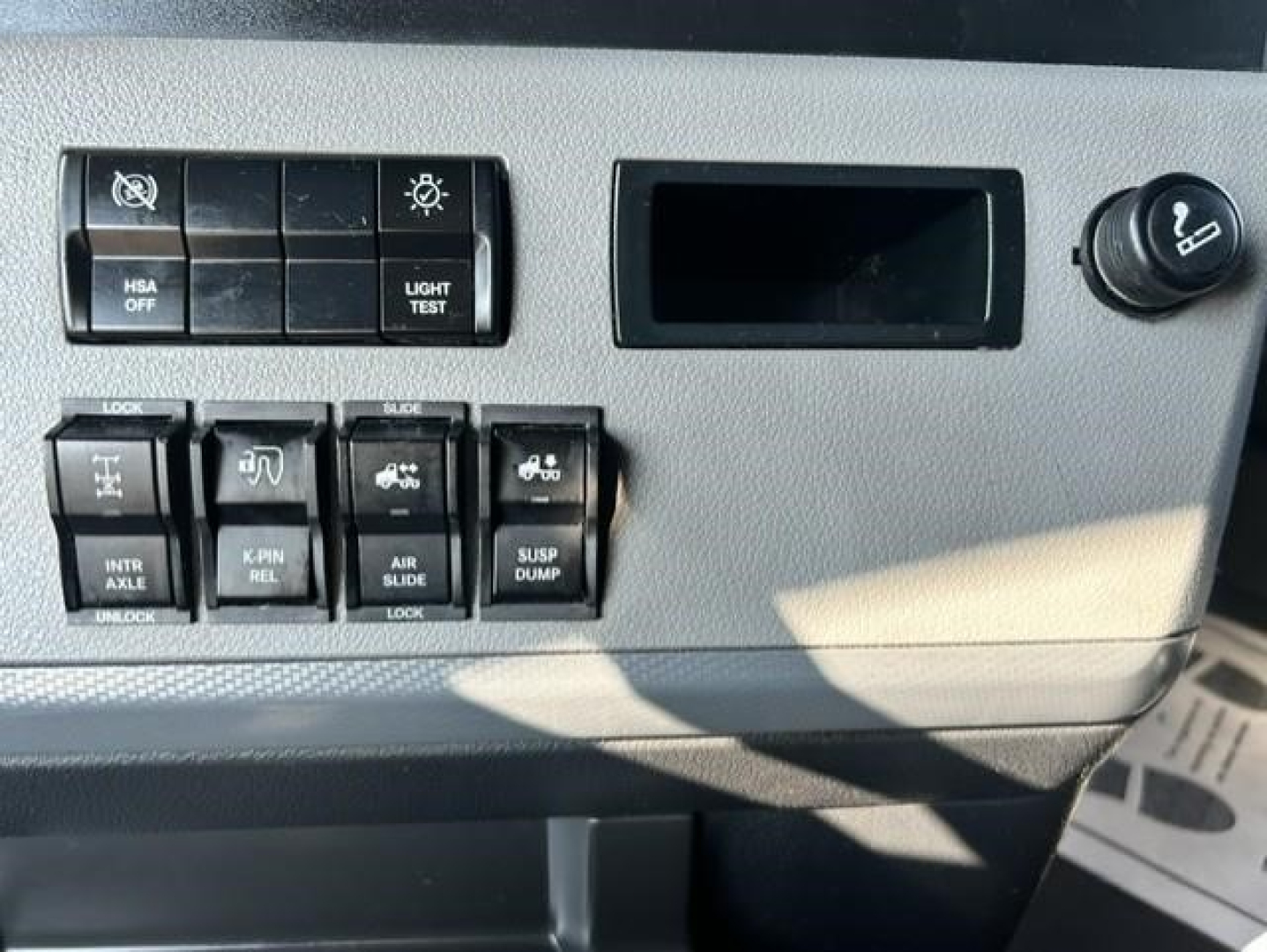 Cab control panel of a Freightliner Cascadia showing switches and knobs with labels such as 'HSA OFF', 'LIGHT TEST', 'INT AXLE LOCK', 'PWR REL', 'AIR SLIDE', and 'SUSP DUMP'.