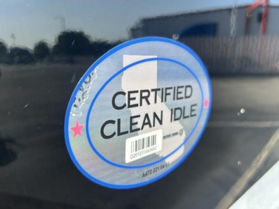 Circular CERTIFIED CLEAN IDLE sticker on a dark surface of a Freightliner Cascadia truck badge. Text includes CERTIFIED CLEAN IDLE with barcode and sticker details.