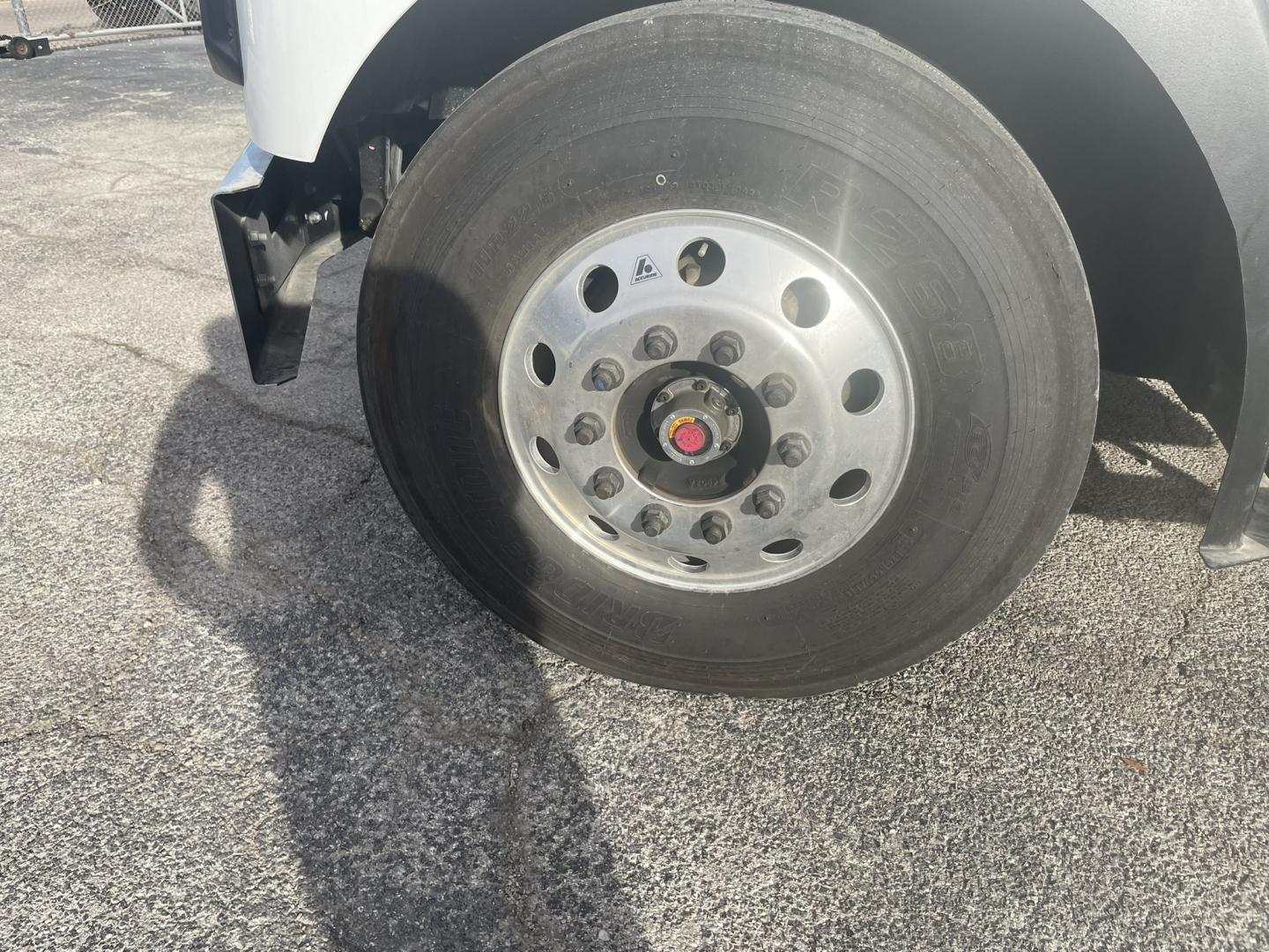 Truck front wheel and hub showing polished steel rim with multiple lug nuts; tire mounted on white body of Mack MD7 refuse truck.