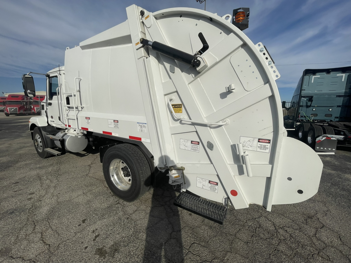 Rear view of a white 2026 Mack MD7 refuse truck showing rear compactor and hydraulic lift mechanism with safety decals and steps.