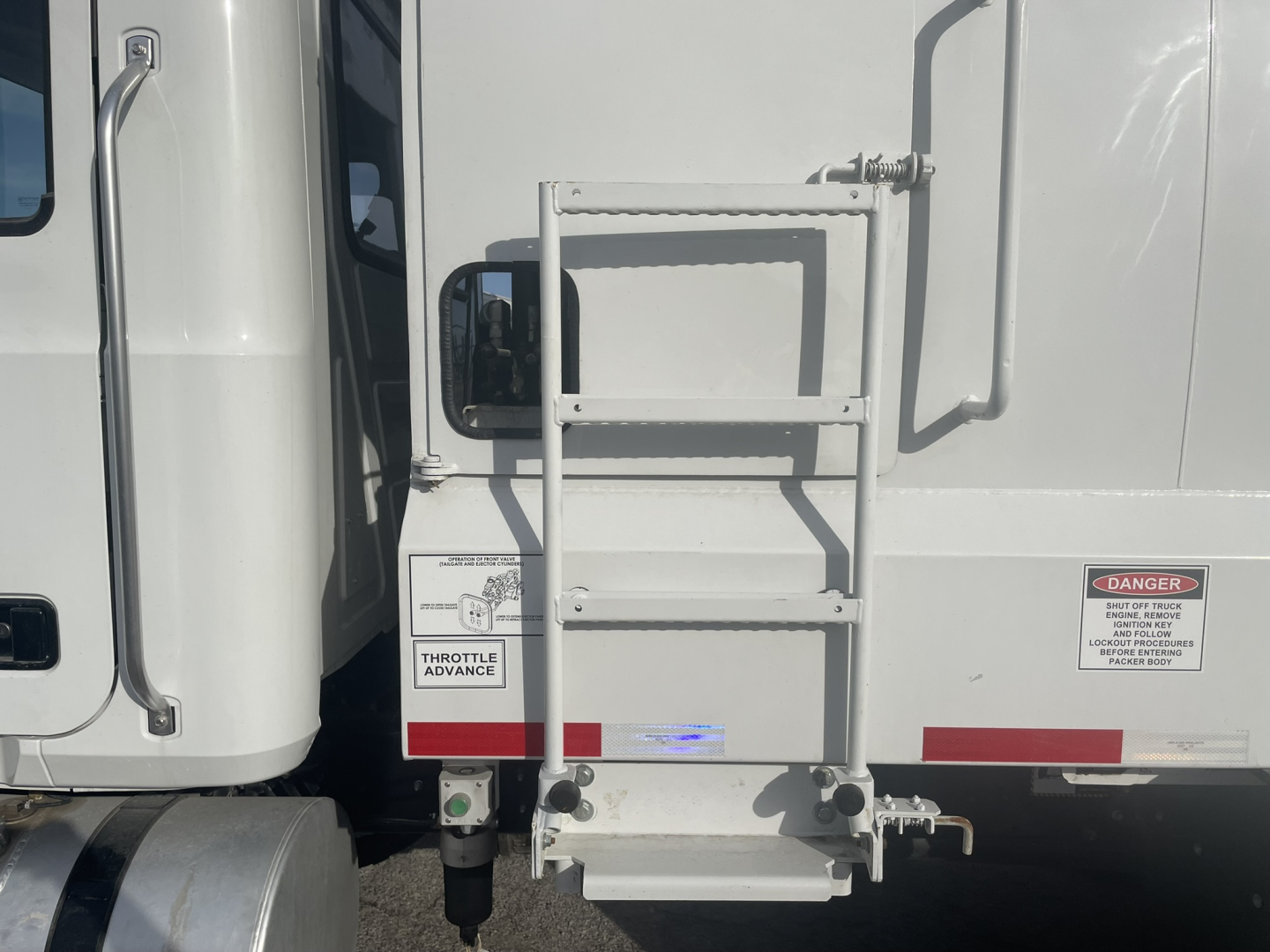 Mack MD7 rear truck view with side ladder, rear bumper, and warning labels. Visible text includes 'DANGER' sticker and 'THROTTLE ADVANCE' label near door area.