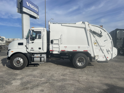 White 2026 Mack MD7 refuse truck with rear compactor body, side view showing cab and dumpster, Volvo Trucks sign in background.