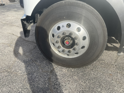 Truck front wheel and hub showing polished steel rim with multiple lug nuts; tire mounted on white body of Mack MD7 refuse truck.
