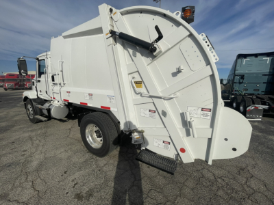 Rear view of a white 2026 Mack MD7 refuse truck showing rear compactor and hydraulic lift mechanism with safety decals and steps.