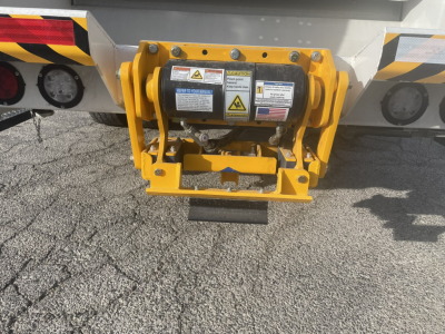 Rear view of a yellow hydraulic air-hook style device attached to a Mack MD7 refuse truck, with warning labels and bolts visible.