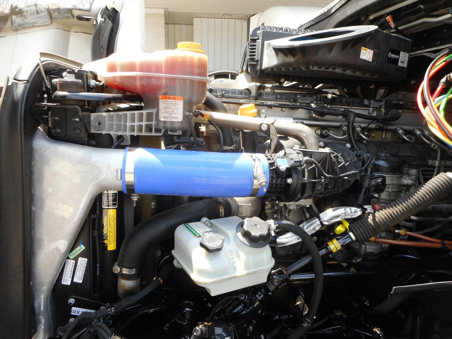 Freightliner Cascadia 126 engine bay showing coolant reservoir, blue silicone intercooler hose, brake reservoir, and various engine components.