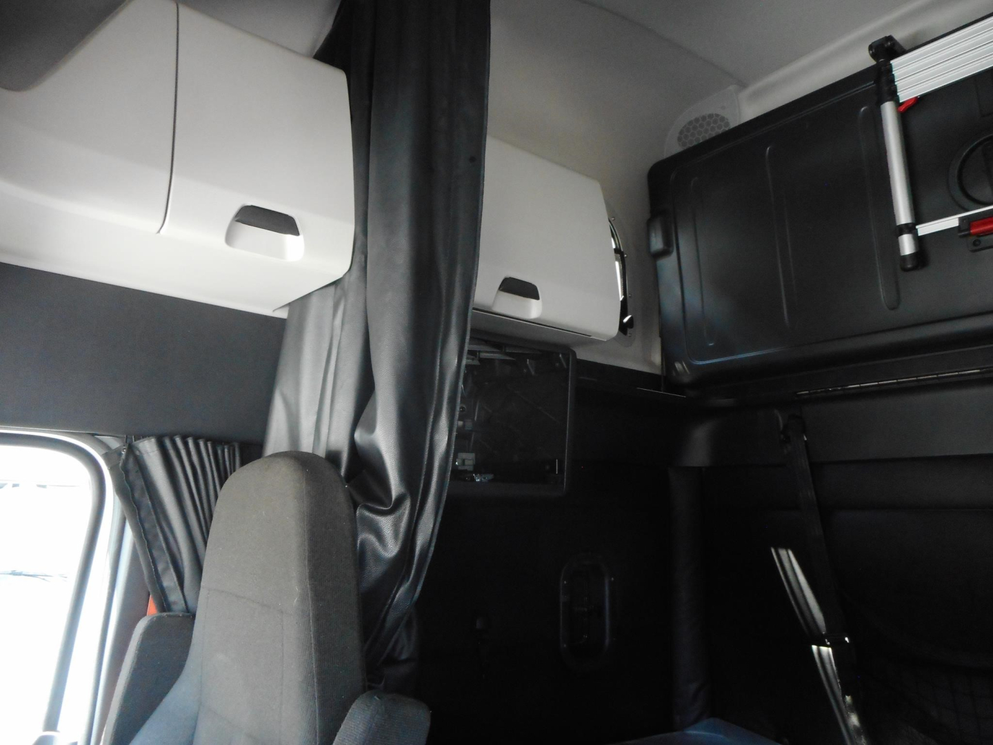 Truck cab interior showing storage compartments and sleeper area in a Freightliner Cascadia with overhead cabinets and a curtain.