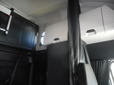 Truck cab interior showing sleeper area with white storage cabinets and black seatbelt and seat; partial view of window and overhead storage.