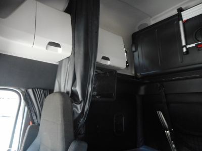 Truck cab interior showing storage compartments and sleeper area in a Freightliner Cascadia with overhead cabinets and a curtain.