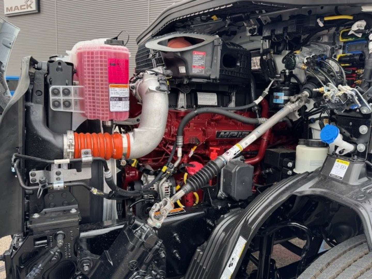 Mack AN64T engine bay showing coolant reservoir, pink coolant overflow tank, red engine block, hoses, and various components.