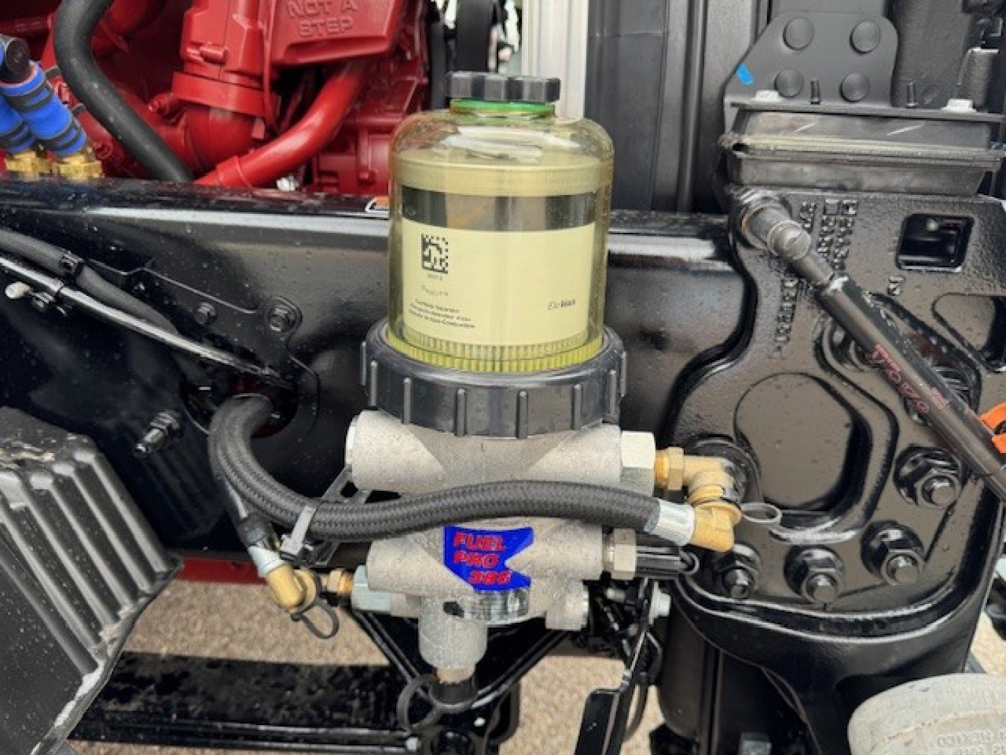 Truck engine bay close-up showing a vertical reservoir with yellowish fluid and a gray pump assembly labeled with blue sticker; surrounding hoses and metal components visible.