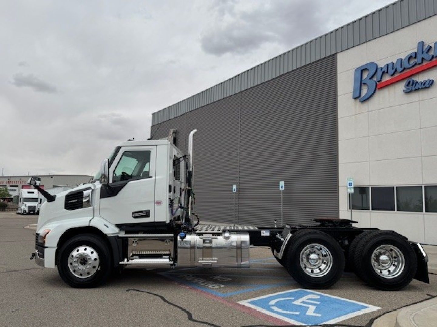 Truck chassis view with a white Mack AN64T cab, side exhaust, and dual rear axles, parked in front of a dealership building.