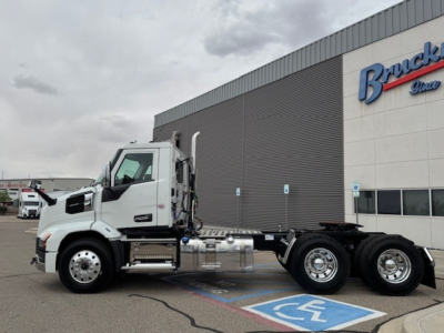 Truck chassis view with a white Mack AN64T cab, side exhaust, and dual rear axles, parked in front of a dealership building.