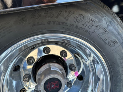Tire and wheel assembly of a Doepker trailer axle showing a large alloy wheel, lug nuts, and a hub with a small label; tread visible on the tire with embossed markings.
