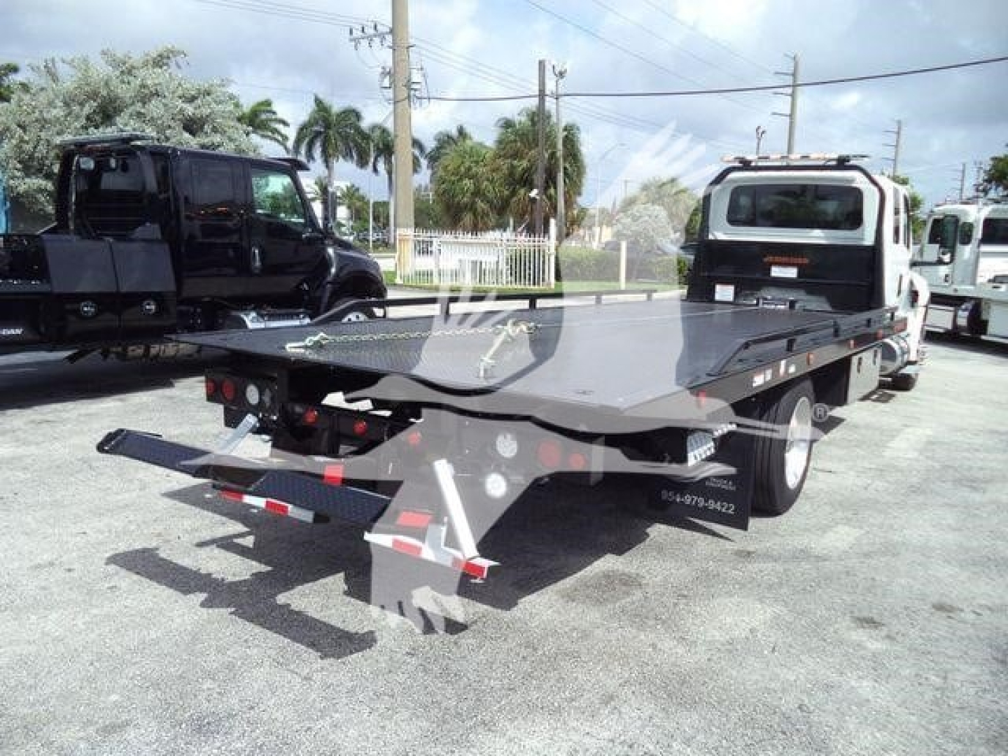 Flatbed tow truck in a dealership lot showing black steel carrier with ramp and rear hitch, International MV rollback style bed, warning lights, and dual rear wheels.