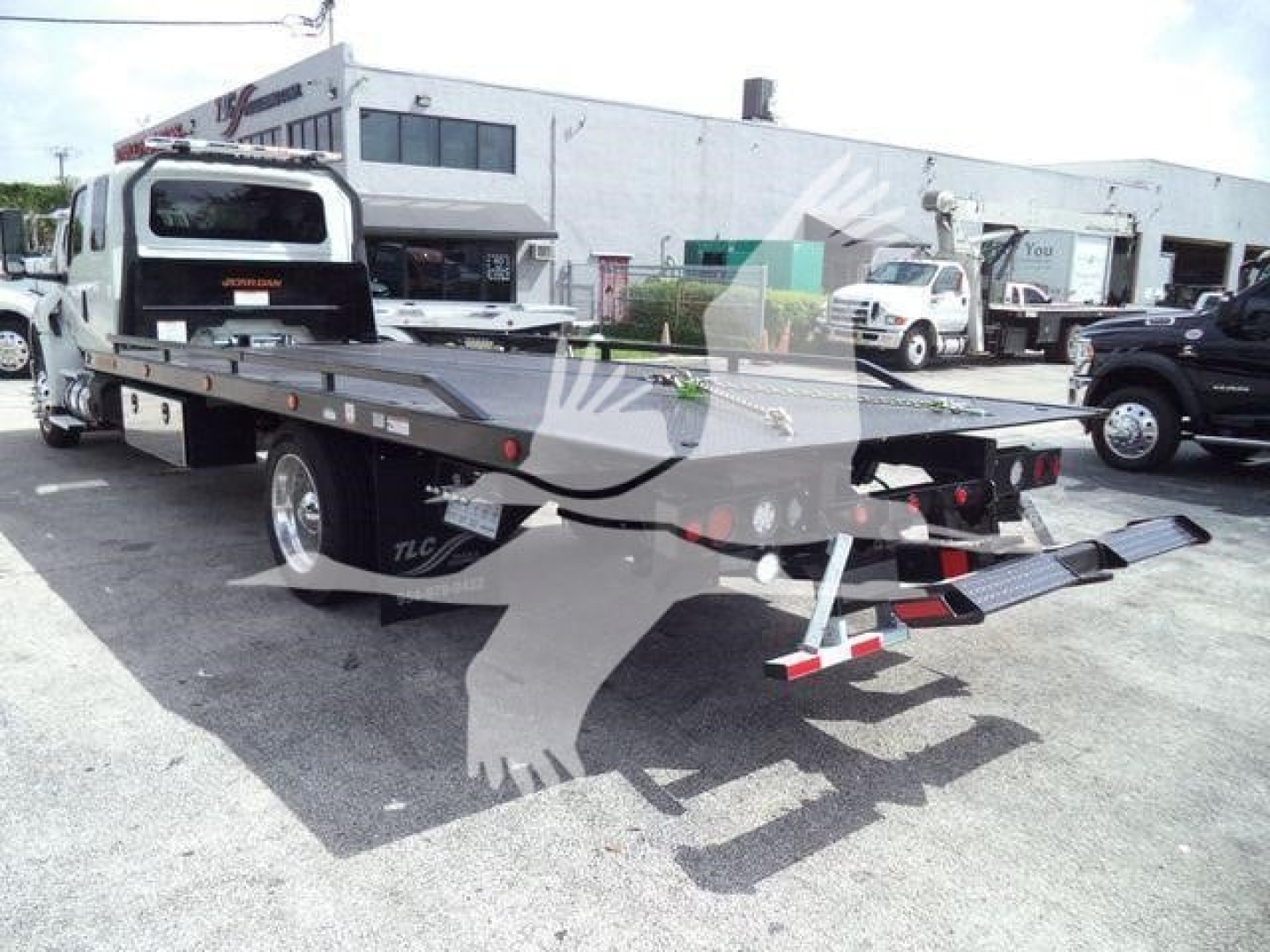 Flatbed tow truck trailer in dealership lot with rear loading ramps and tail lights visible.