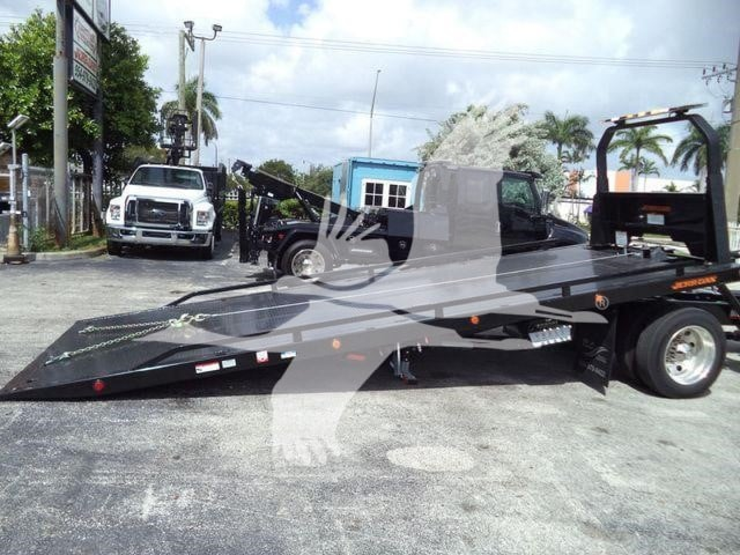 Flatbed tow truck trailer in a yard with another white truck in background; "MV" and equipment markings are visible on trailer.