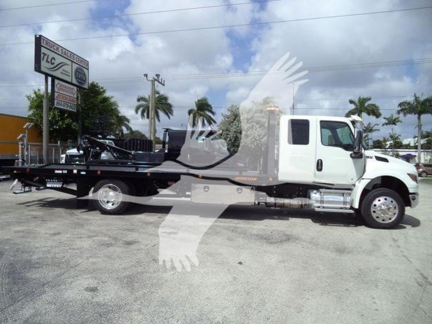 Truck with a rollback bed and crane in a parking lot, white cab and flatbed, International MV model.