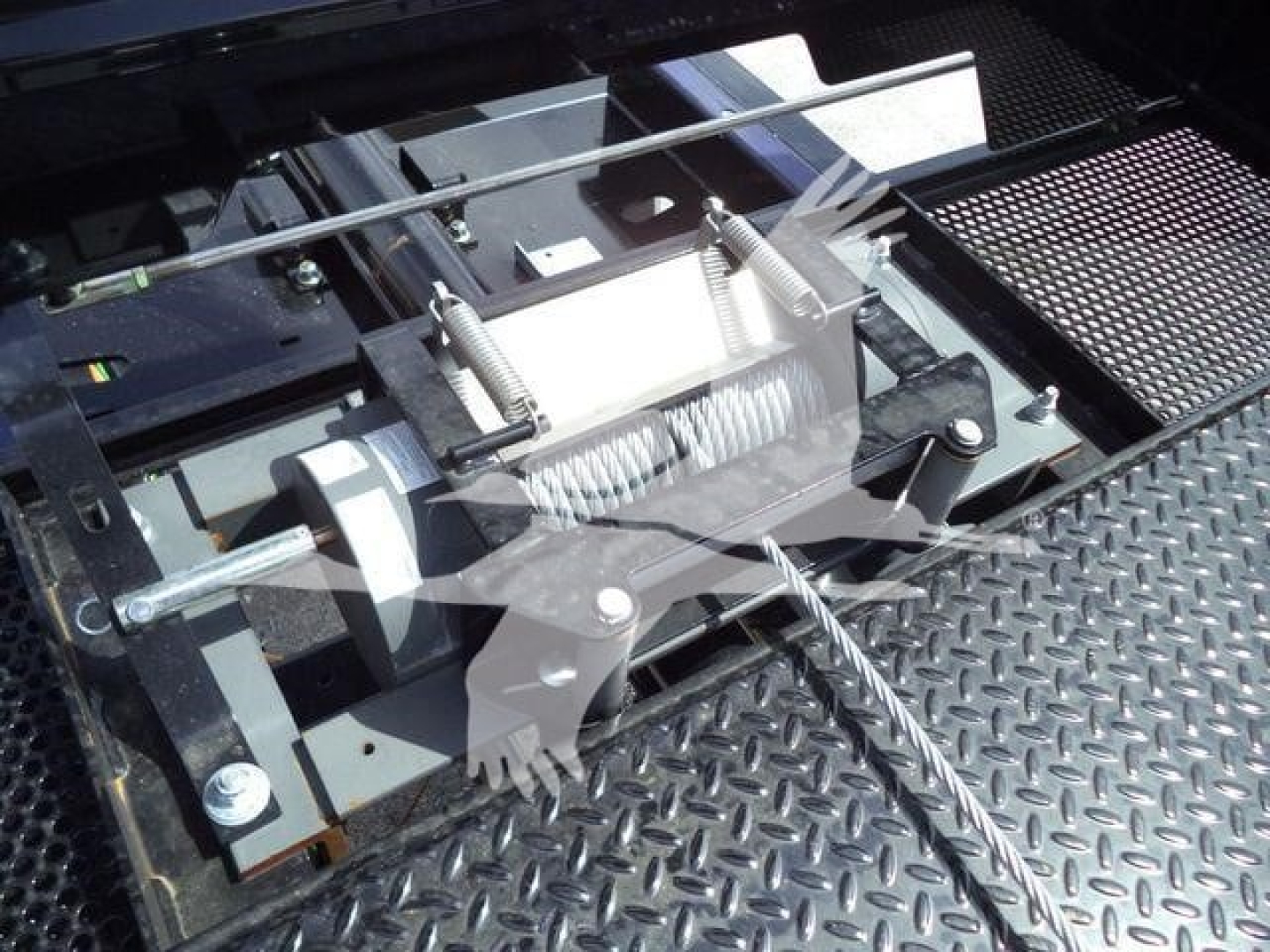 Trailer winch and mounting hardware with steel cable and threaded rods on a diamond plate surface. Visible components include a winch drum, spring-loaded tensioner, and mounting brackets.