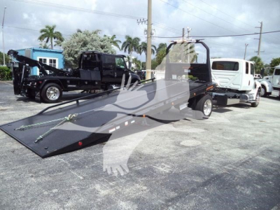 Flatbed tow truck with black ramp bed and white tow truck attached, in a dealership lot.